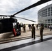 A U.S. Army UH-60 Black Hawk helicopter is parked on a tarmac adjacent to a modern curved glass building, likely a terminal or aviation facility. Military personnel in camouflage uniforms and civilians are gathered around the aircraft, suggesting a demonstration, open house, or familiarization event. The scene appears to be at a National Guard or military airfield facility.