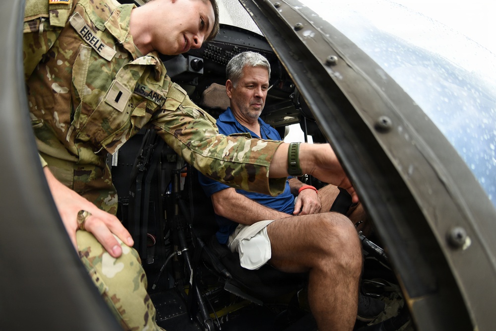 A U.S. Army soldier with the name tape 'EISELE' is assisting a civilian man seated inside what appears to be a military helicopter cockpit. The soldier, wearing Operational Camouflage Pattern (OCP) uniform with 'U.S. ARMY' tape visible, appears to be orienting or briefing the civilian on the aircraft's interior. The setting suggests a public demonstration, airshow, or familiarization event.