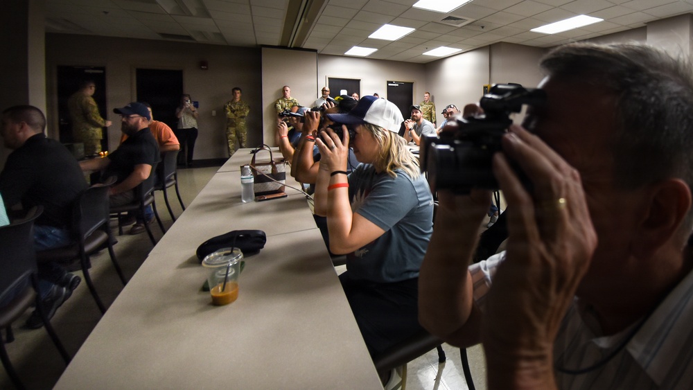 A press briefing or media event is taking place in what appears to be a conference room at a military installation. Multiple photographers and journalists are capturing images, while military personnel in camouflage uniforms stand in the background. The scene suggests an official media access event or press conference.