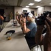 A press briefing or media event is taking place in what appears to be a conference room at a military installation. Multiple photographers and journalists are capturing images, while military personnel in camouflage uniforms stand in the background. The scene suggests an official media access event or press conference.