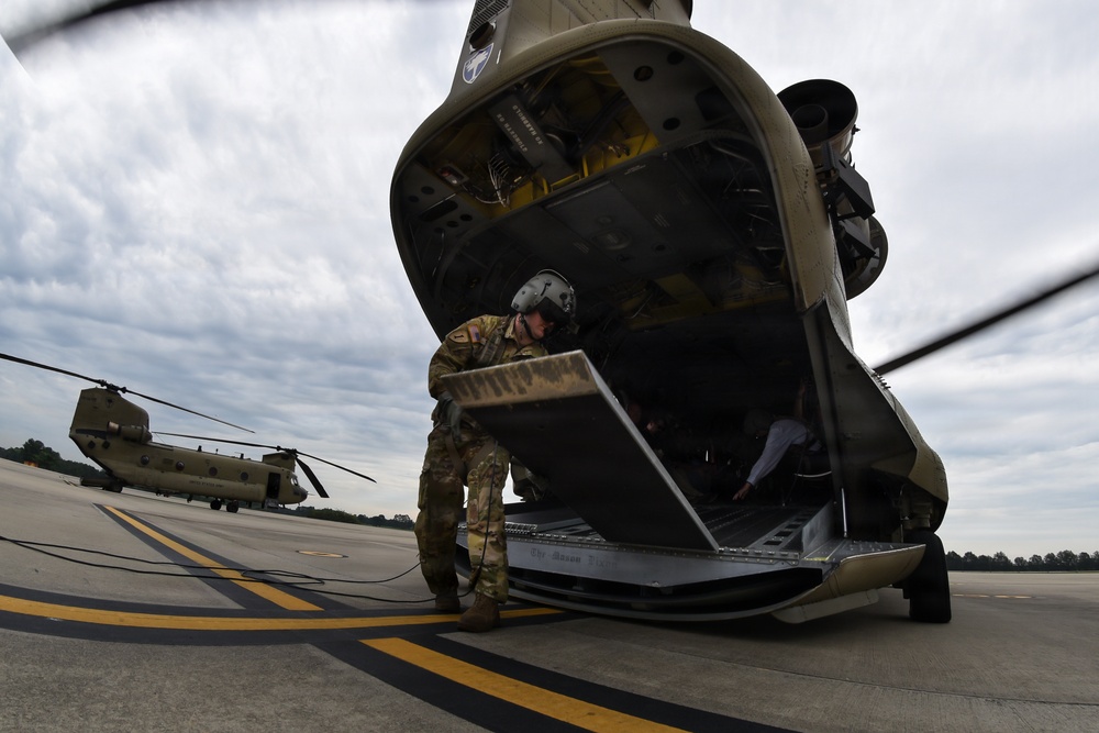 A military service member in combat uniform and flight helmet is operating the rear loading ramp of a CH-47 Chinook helicopter on a tarmac. The wide-angle fisheye lens creates a dramatic perspective of the large tandem-rotor helicopter's aft section. A second CH-47 Chinook helicopter is visible in the background on the same flight line.