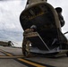A military service member in combat uniform and flight helmet is operating the rear loading ramp of a CH-47 Chinook helicopter on a tarmac. The wide-angle fisheye lens creates a dramatic perspective of the large tandem-rotor helicopter's aft section. A second CH-47 Chinook helicopter is visible in the background on the same flight line.