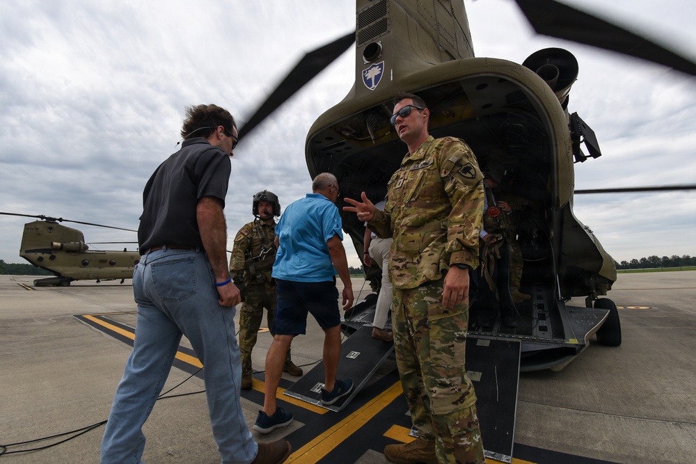 A U.S. Army soldier in OCP (Operational Camouflage Pattern) uniform is briefing or speaking to civilian visitors near the rear ramp of a CH-47 Chinook helicopter on a military airfield. A second soldier in flight gear is visible in the background, along with at least two civilians. A second Chinook helicopter is visible in the background on the tarmac.