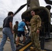 A U.S. Army soldier in OCP (Operational Camouflage Pattern) uniform is briefing or speaking to civilian visitors near the rear ramp of a CH-47 Chinook helicopter on a military airfield. A second soldier in flight gear is visible in the background, along with at least two civilians. A second Chinook helicopter is visible in the background on the tarmac.