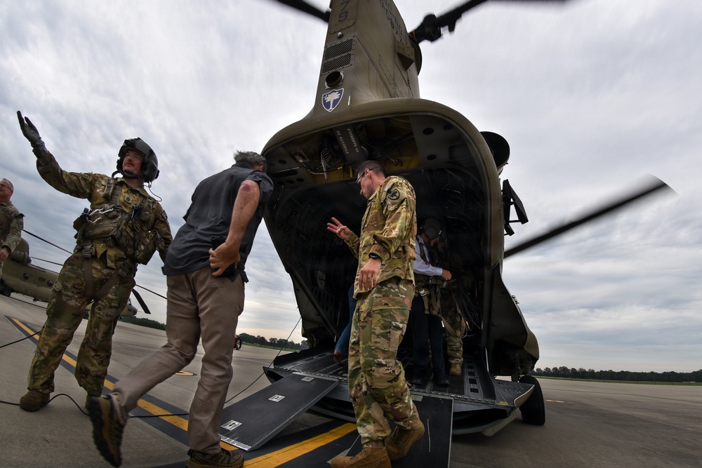 Military personnel in OCP (Operational Camouflage Pattern) uniforms are directing civilians onto or off of a CH-47 Chinook helicopter on a tarmac. The scene appears to be a boarding or orientation event, with soldiers gesturing to guide individuals toward the rear ramp of the aircraft. The overcast sky and open tarmac suggest an outdoor military airfield setting.