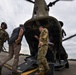 Military personnel in OCP (Operational Camouflage Pattern) uniforms are directing civilians onto or off of a CH-47 Chinook helicopter on a tarmac. The scene appears to be a boarding or orientation event, with soldiers gesturing to guide individuals toward the rear ramp of the aircraft. The overcast sky and open tarmac suggest an outdoor military airfield setting.