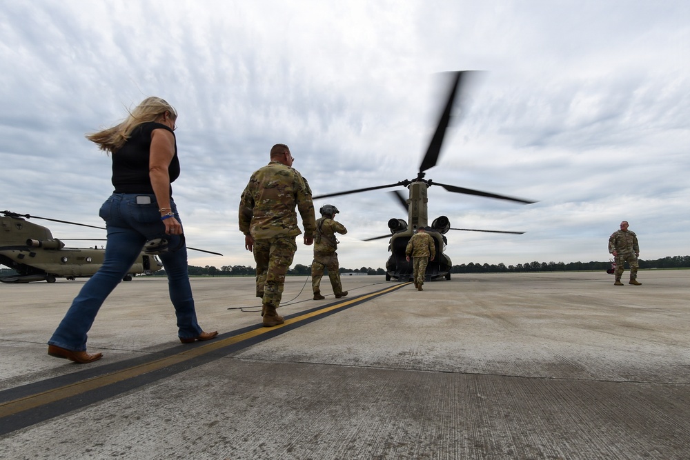 Military personnel in camouflage uniforms and one civilian woman are walking across a tarmac toward a CH-47 Chinook tandem-rotor helicopter with its rotors spinning. A second Chinook is visible in the background to the left. The scene appears to depict a pre-boarding or operational activity at a military airfield.