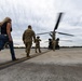 Military personnel in camouflage uniforms and one civilian woman are walking across a tarmac toward a CH-47 Chinook tandem-rotor helicopter with its rotors spinning. A second Chinook is visible in the background to the left. The scene appears to depict a pre-boarding or operational activity at a military airfield.