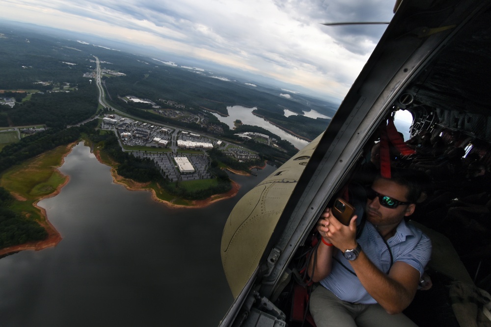 A man wearing sunglasses and a blue shirt is leaning out of an open helicopter door, photographing the landscape below with a smartphone. The aerial view reveals a suburban area with a reservoir or lake, commercial buildings, parking lots, and a highway cutting through forested terrain. The overcast sky and wide-angle perspective suggest this is a media or reconnaissance flight.