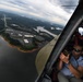 A man wearing sunglasses and a blue shirt is leaning out of an open helicopter door, photographing the landscape below with a smartphone. The aerial view reveals a suburban area with a reservoir or lake, commercial buildings, parking lots, and a highway cutting through forested terrain. The overcast sky and wide-angle perspective suggest this is a media or reconnaissance flight.