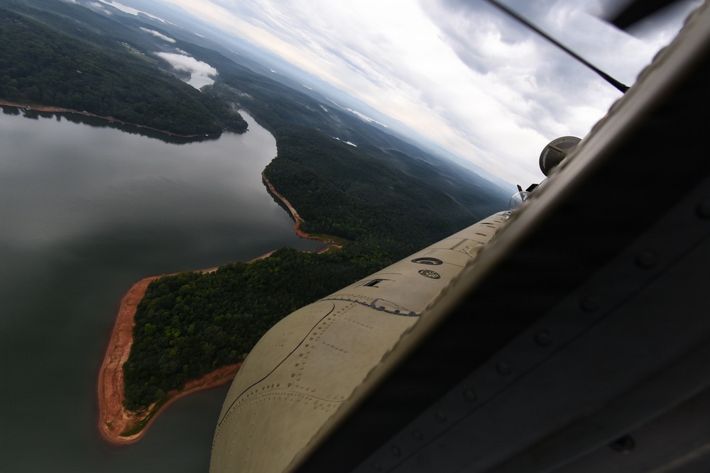 An aerial photograph taken from inside a small propeller-driven aircraft, showing a bird's-eye view of a large reservoir or lake surrounded by dense green forest. The aircraft wing and engine are visible in the foreground, with red-clay exposed shorelines and a winding waterway visible below. Overcast skies with scattered clouds extend to the horizon over a heavily forested landscape.