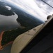 An aerial photograph taken from inside a small propeller-driven aircraft, showing a bird's-eye view of a large reservoir or lake surrounded by dense green forest. The aircraft wing and engine are visible in the foreground, with red-clay exposed shorelines and a winding waterway visible below. Overcast skies with scattered clouds extend to the horizon over a heavily forested landscape.