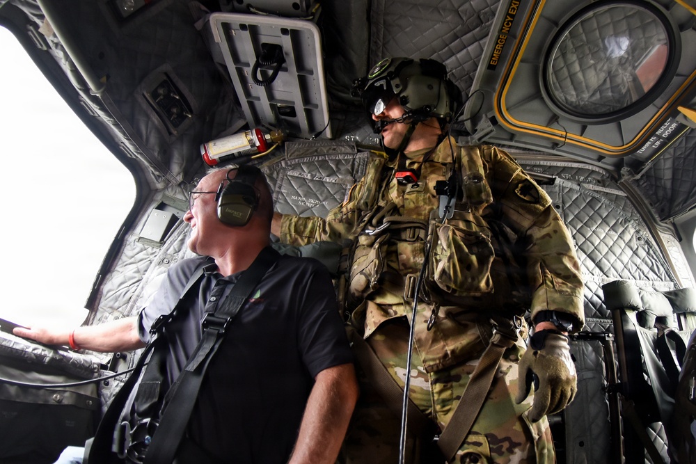 The image shows the interior of a military helicopter with two individuals visible. One person is wearing full military combat gear including a flight helmet with night vision mount, multicam camouflage uniform, tactical vest, and gloves. The other individual is a civilian wearing a black polo shirt and aviation headset, appearing to be a passenger or observer.