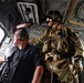 The image shows the interior of a military helicopter with two individuals visible. One person is wearing full military combat gear including a flight helmet with night vision mount, multicam camouflage uniform, tactical vest, and gloves. The other individual is a civilian wearing a black polo shirt and aviation headset, appearing to be a passenger or observer.