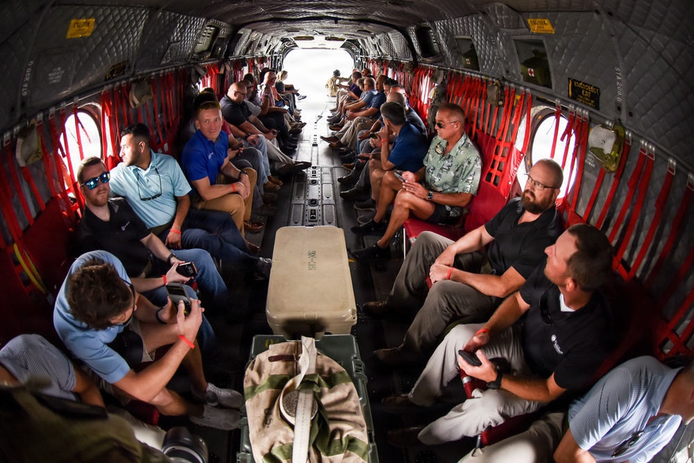 The image shows the interior of a large military helicopter, likely a CH-47 Chinook, filled with approximately 20-25 civilian passengers seated along both sides of the fuselage. The passengers appear to be civilians or officials on an orientation or familiarization flight, dressed in casual and business-casual attire. A fisheye lens was used to capture the full length of the cargo bay.