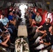 The image shows the interior of a large military helicopter, likely a CH-47 Chinook, filled with approximately 20-25 civilian passengers seated along both sides of the fuselage. The passengers appear to be civilians or officials on an orientation or familiarization flight, dressed in casual and business-casual attire. A fisheye lens was used to capture the full length of the cargo bay.