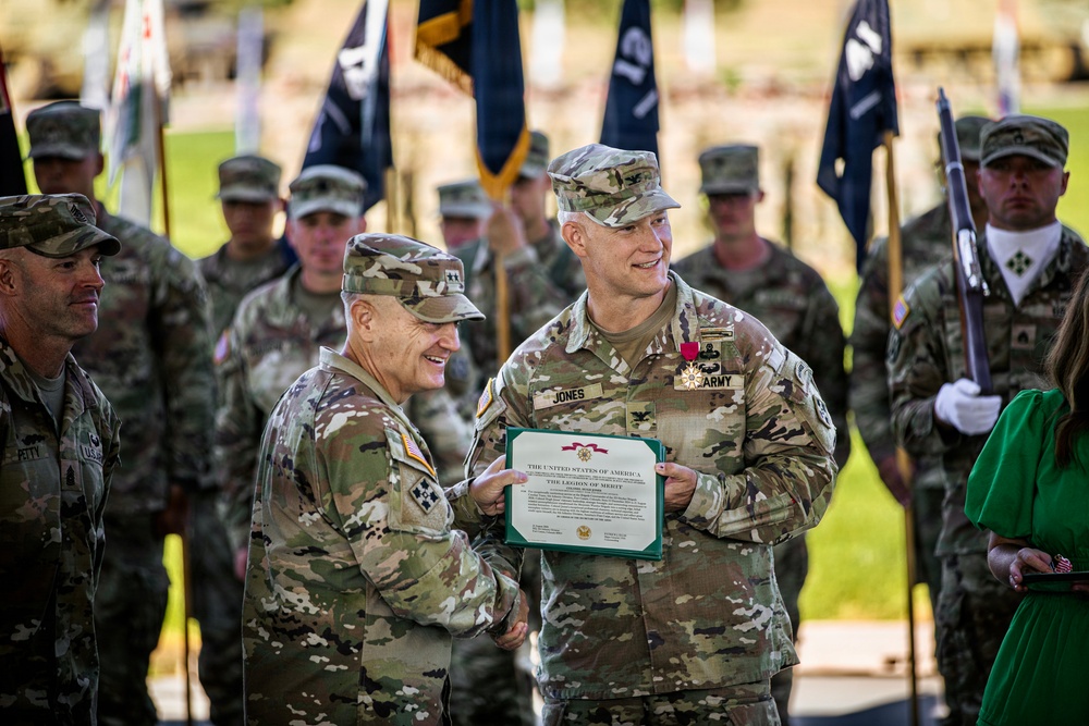 A U.S. Army military ceremony in which a soldier named Jones is receiving the Legion of Merit award. Two officers are jointly holding the award certificate while smiling, with the recipient also wearing the Legion of Merit medal on his chest. A formation of soldiers and color guard with military flags stands in the background.