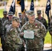 A U.S. Army military ceremony in which a soldier named Jones is receiving the Legion of Merit award. Two officers are jointly holding the award certificate while smiling, with the recipient also wearing the Legion of Merit medal on his chest. A formation of soldiers and color guard with military flags stands in the background.