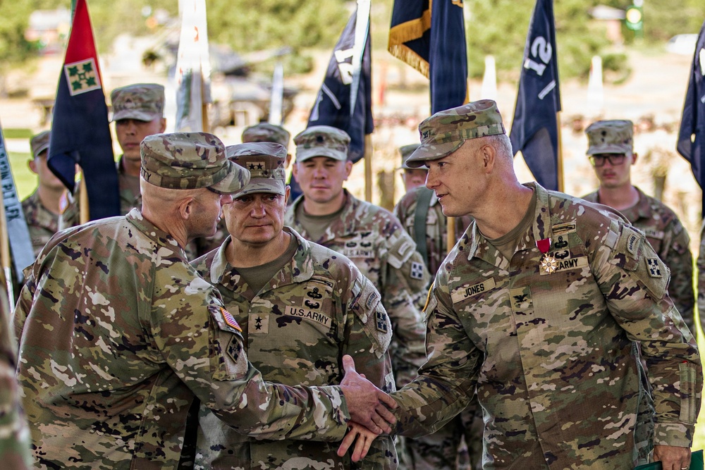 A formal U.S. Army military ceremony is taking place outdoors, featuring senior officers in OCP (Operational Camouflage Pattern) uniforms. Two officers are shaking hands in the foreground, with one identified by nametape as 'JONES' wearing a medal (appears to be a Meritorious Service Medal or Legion of Merit). Multiple unit guidons and color guard personnel are visible in the background, suggesting a change of command or awards ceremony.