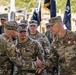 A formal U.S. Army military ceremony is taking place outdoors, featuring senior officers in OCP (Operational Camouflage Pattern) uniforms. Two officers are shaking hands in the foreground, with one identified by nametape as 'JONES' wearing a medal (appears to be a Meritorious Service Medal or Legion of Merit). Multiple unit guidons and color guard personnel are visible in the background, suggesting a change of command or awards ceremony.