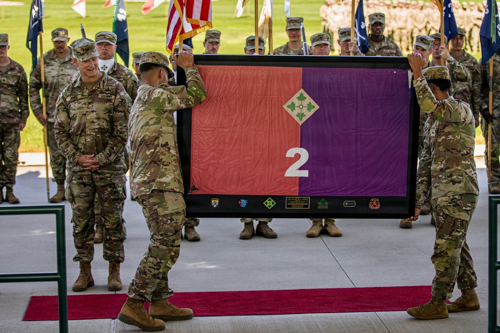 A U.S. Army military ceremony is taking place outdoors, likely a change of command or unit activation/inactivation ceremony. Two soldiers are holding up a framed flag displaying the number '2' on a red and purple background with a 4th Infantry Division insignia (four-leaf clover diamond). A formation of soldiers stands in the background alongside multiple unit guidons and the American flag.