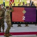 A U.S. Army military ceremony is taking place outdoors, likely a change of command or unit activation/inactivation ceremony. Two soldiers are holding up a framed flag displaying the number '2' on a red and purple background with a 4th Infantry Division insignia (four-leaf clover diamond). A formation of soldiers stands in the background alongside multiple unit guidons and the American flag.