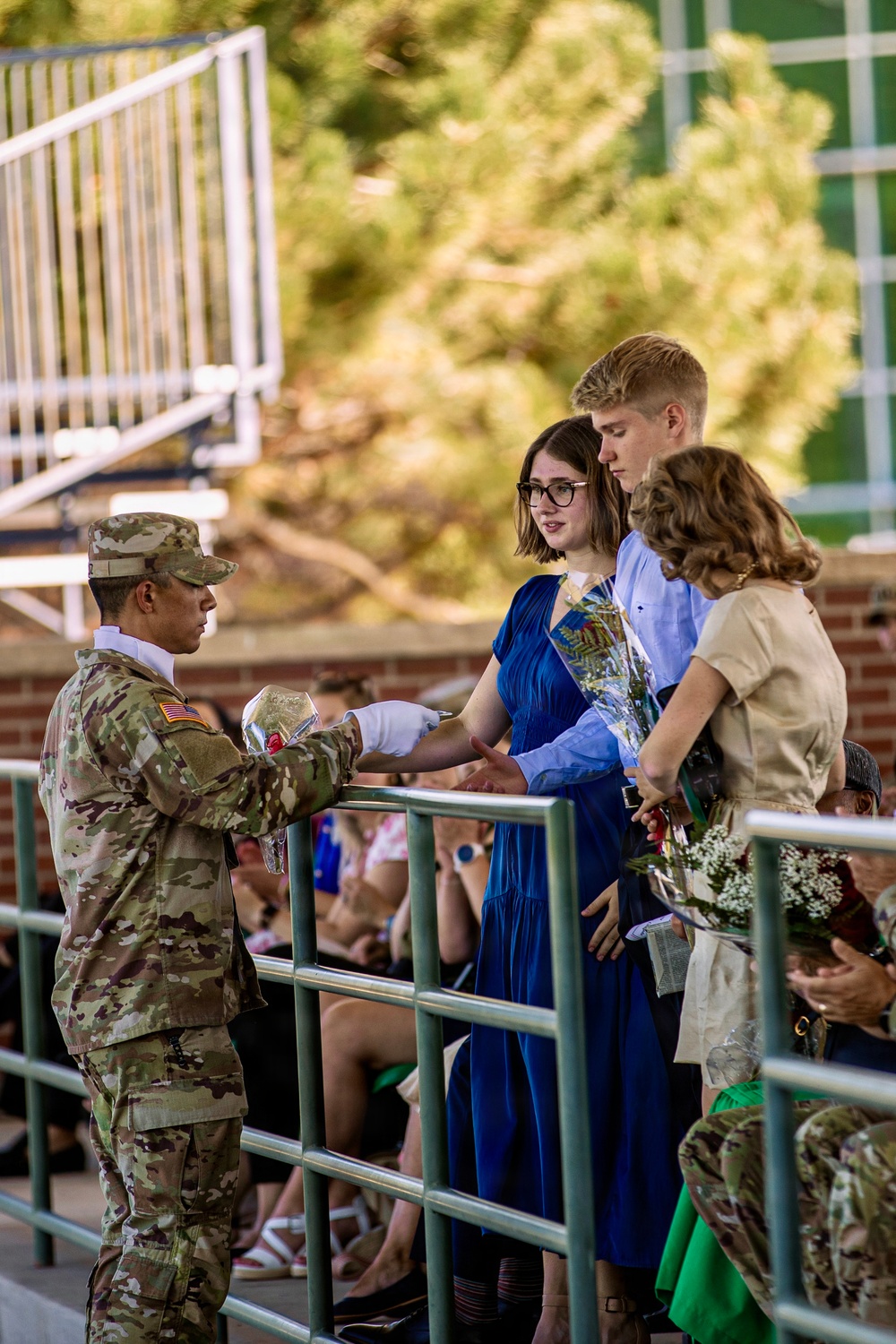 A U.S. Army soldier in OCP (Operational Camouflage Pattern) uniform is presenting or handing an item to a young woman in a blue dress across a metal railing, accompanied by two other civilians. The setting appears to be an outdoor military ceremony or commissioning event, with a seated audience visible in the background. The soldier is wearing white gloves, suggesting a formal ceremonial occasion.