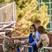 A U.S. Army soldier in OCP (Operational Camouflage Pattern) uniform is presenting or handing an item to a young woman in a blue dress across a metal railing, accompanied by two other civilians. The setting appears to be an outdoor military ceremony or commissioning event, with a seated audience visible in the background. The soldier is wearing white gloves, suggesting a formal ceremonial occasion.