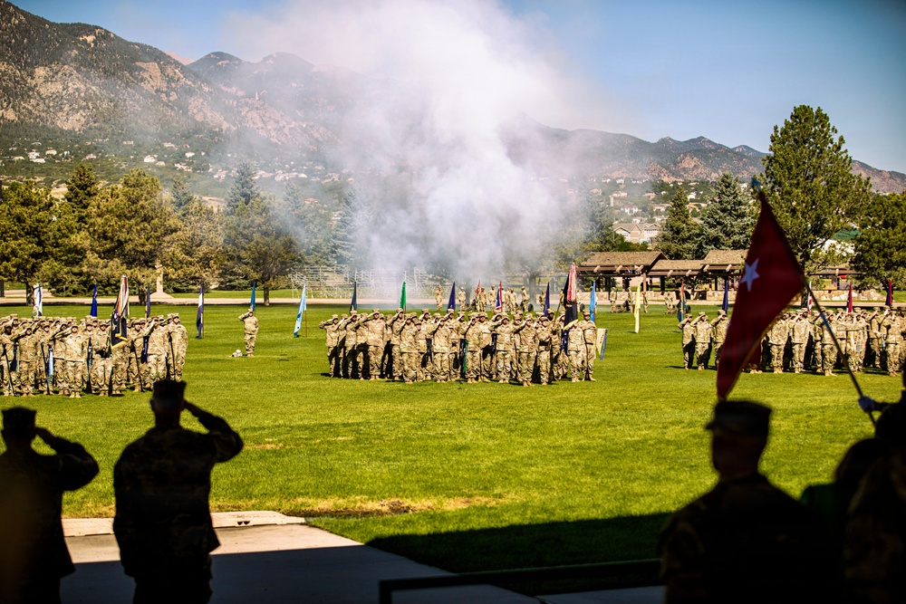 A formal military ceremony is taking place on a large green parade field with multiple units in formation. Soldiers in camouflage uniforms are saluting while color guards hold various unit guidons and flags, including a red general officer flag visible in the foreground. A cannon or artillery piece has just been fired, producing a large smoke cloud above the field, with a mountain range visible in the background.