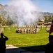 A formal military ceremony is taking place on a large green parade field with multiple units in formation. Soldiers in camouflage uniforms are saluting while color guards hold various unit guidons and flags, including a red general officer flag visible in the foreground. A cannon or artillery piece has just been fired, producing a large smoke cloud above the field, with a mountain range visible in the background.