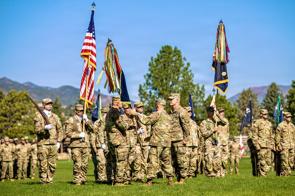 A formal U.S. Army military ceremony, likely a change of command, is taking place on a green parade field with mountains and pine trees visible in the background. Several soldiers in OCP (Operational Camouflage Pattern) uniforms are participating in a guidon or colors transfer at the center of the formation. Rows of soldiers stand at attention in the background while color bearers hold multiple flags including the U.S. national flag and unit guidons.