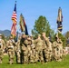 A formal U.S. Army military ceremony, likely a change of command, is taking place on a green parade field with mountains and pine trees visible in the background. Several soldiers in OCP (Operational Camouflage Pattern) uniforms are participating in a guidon or colors transfer at the center of the formation. Rows of soldiers stand at attention in the background while color bearers hold multiple flags including the U.S. national flag and unit guidons.