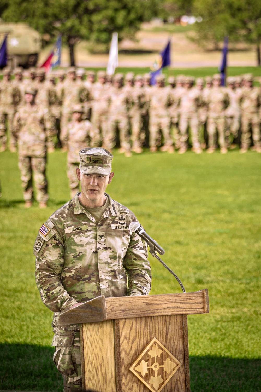 A U.S. Army Colonel named Jones is speaking at a podium during a formal military ceremony on a grass field. The podium bears the insignia of the 4th Infantry Division. A large formation of soldiers in OCP (Operational Camouflage Pattern) uniforms stands at attention in the background, with multiple unit guidons/flags visible.