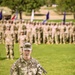 A U.S. Army Colonel named Jones is speaking at a podium during a formal military ceremony on a grass field. The podium bears the insignia of the 4th Infantry Division. A large formation of soldiers in OCP (Operational Camouflage Pattern) uniforms stands at attention in the background, with multiple unit guidons/flags visible.