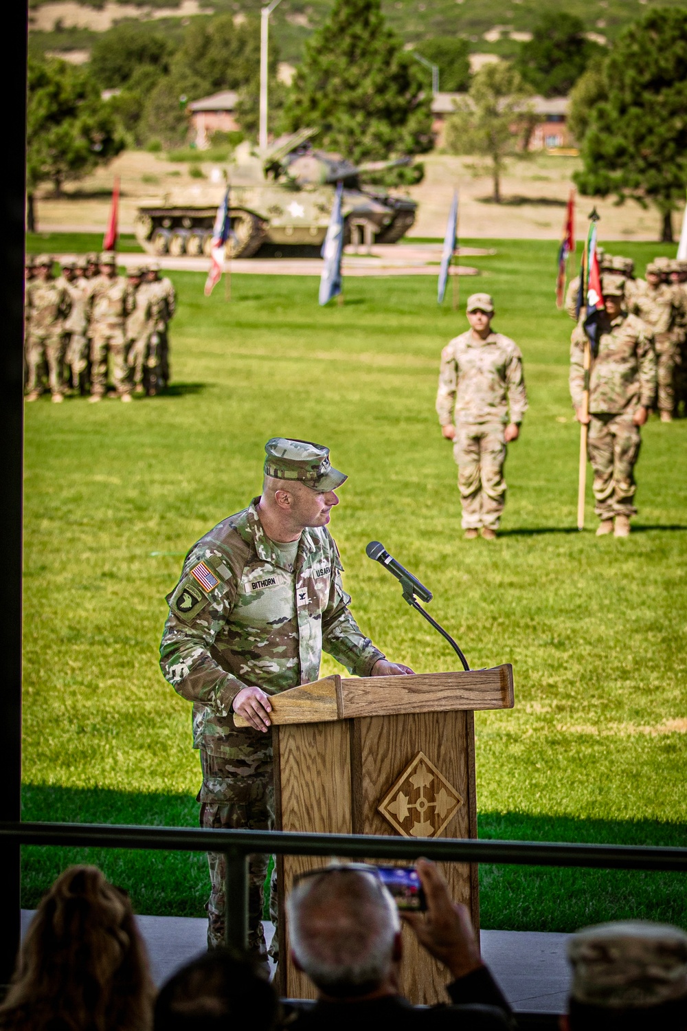 A U.S. Army officer named Bithorn, wearing OCP (Operational Camouflage Pattern) uniform with a 101st Airborne Division patch, speaks at a wooden podium bearing the 4th Infantry Division insignia. The ceremony appears to be a formal military event, likely a change of command or activation ceremony, held outdoors on a parade field. Formations of soldiers, military vehicles, and color guard personnel are visible in the background.