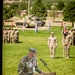 A U.S. Army officer named Bithorn, wearing OCP (Operational Camouflage Pattern) uniform with a 101st Airborne Division patch, speaks at a wooden podium bearing the 4th Infantry Division insignia. The ceremony appears to be a formal military event, likely a change of command or activation ceremony, held outdoors on a parade field. Formations of soldiers, military vehicles, and color guard personnel are visible in the background.