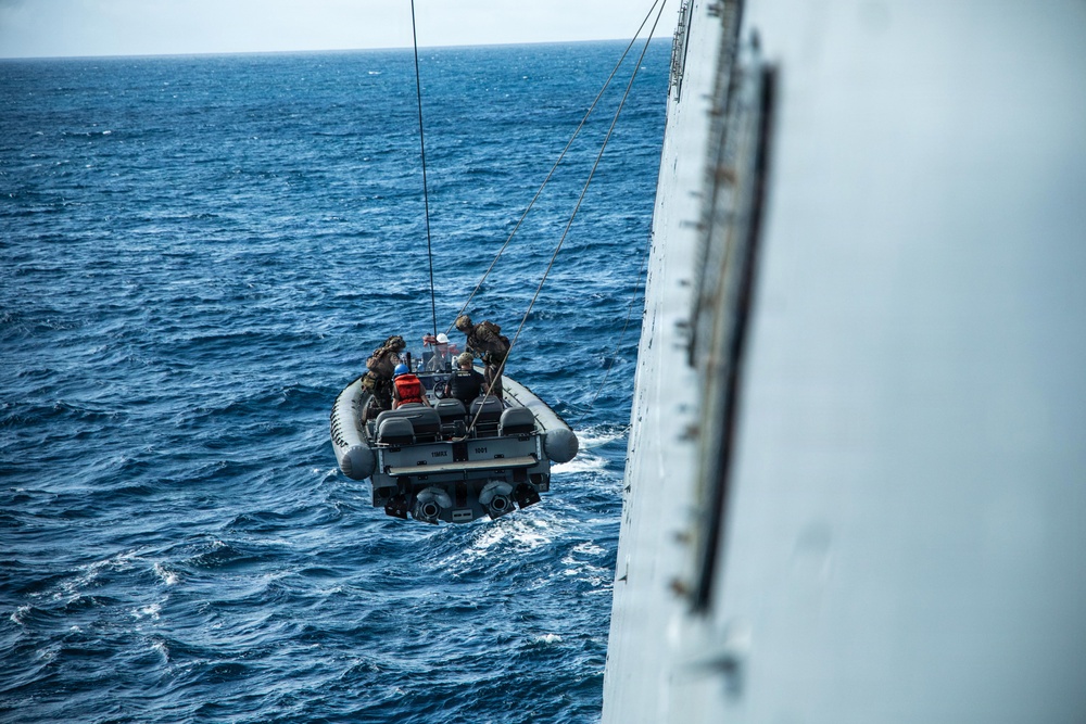 A rigid inflatable boat (RIB) is being hoisted or lowered alongside a large naval vessel using davit lines over open ocean. Several personnel in military/tactical gear are aboard the small craft during the operation. The image is taken from an elevated angle on the ship's hull, showing the boat partially suspended above choppy blue water.