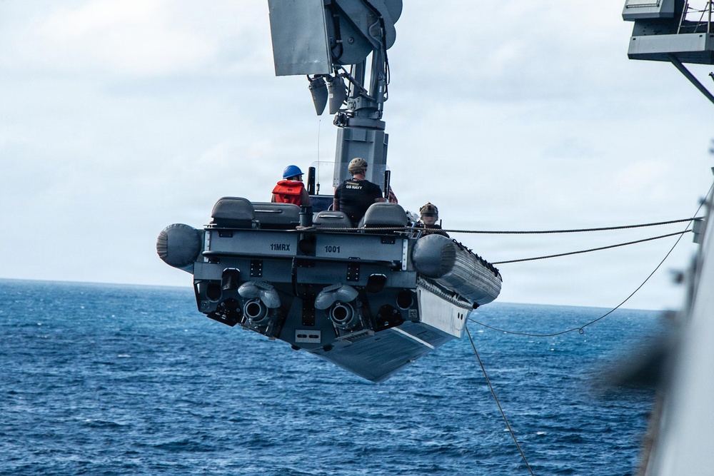 A rigid-hull inflatable boat (RHIB) designated '11MRX 1001' is being hoisted by a crane davit from a US Navy vessel over open ocean. Three personnel are visible aboard the suspended small craft, with one wearing a blue hard hat and another in a US Navy-marked shirt. The operation appears to be a boat launch or recovery evolution conducted at sea.