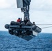 A rigid-hull inflatable boat (RHIB) designated '11MRX 1001' is being hoisted by a crane davit from a US Navy vessel over open ocean. Three personnel are visible aboard the suspended small craft, with one wearing a blue hard hat and another in a US Navy-marked shirt. The operation appears to be a boat launch or recovery evolution conducted at sea.