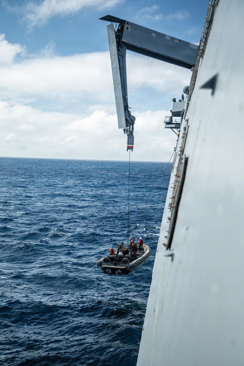 A rigid inflatable boat (RIB) is being hoisted or lowered alongside a large naval vessel using a davit crane system over open ocean. Several personnel are visible aboard the small boat, with at least two wearing high-visibility helmets. The operation appears to be a boat deployment or recovery exercise conducted at sea.