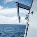 A rigid inflatable boat (RIB) is being hoisted or lowered alongside a large naval vessel using a davit crane system over open ocean. Several personnel are visible aboard the small boat, with at least two wearing high-visibility helmets. The operation appears to be a boat deployment or recovery exercise conducted at sea.