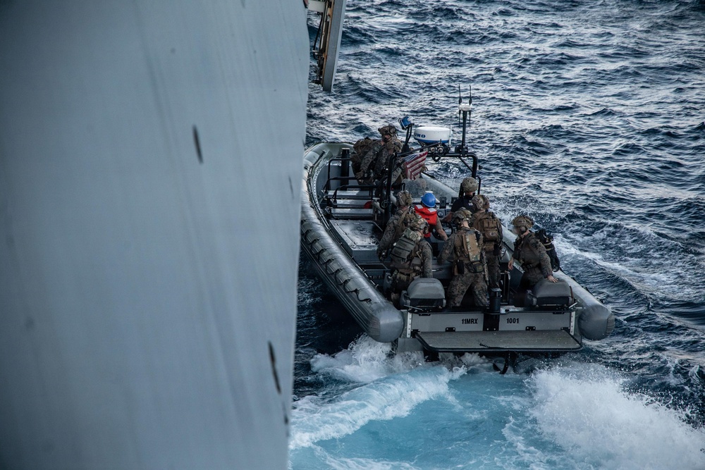 A rigid-hull inflatable boat (RHIB) marked '11MRX 1001' carrying multiple military personnel in combat gear approaches the hull of a large vessel at sea. The personnel appear to be conducting a maritime boarding or visit, board, search, and seizure (VBSS) operation or training exercise. A small American flag is visible on the bow of the RHIB.