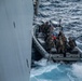 A rigid-hull inflatable boat (RHIB) marked '11MRX 1001' carrying multiple military personnel in combat gear approaches the hull of a large vessel at sea. The personnel appear to be conducting a maritime boarding or visit, board, search, and seizure (VBSS) operation or training exercise. A small American flag is visible on the bow of the RHIB.