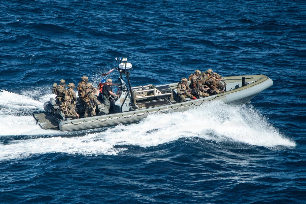 A rigid-hull inflatable boat (RHIB) is underway at high speed on open blue water, carrying multiple personnel in military combat gear and camouflage uniforms. The boat appears to be a military or law enforcement tactical vessel equipped with a center console, navigation/sensor equipment mounted on a mast, and outboard engines. The image is taken from an elevated aerial perspective, likely from a ship or helicopter.