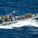 A rigid-hull inflatable boat (RHIB) is underway at high speed on open blue water, carrying multiple personnel in military combat gear and camouflage uniforms. The boat appears to be a military or law enforcement tactical vessel equipped with a center console, navigation/sensor equipment mounted on a mast, and outboard engines. The image is taken from an elevated aerial perspective, likely from a ship or helicopter.
