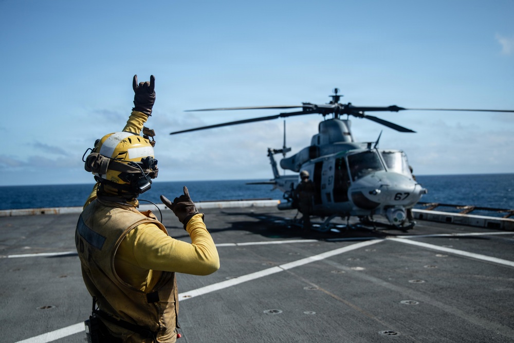 A flight deck crew member in yellow gear is signaling a military helicopter on the deck of a naval vessel at sea. The crew member is using hand signals to communicate with the helicopter crew, a standard procedure during flight deck operations. The helicopter, marked with the number '67', appears to be a U.S. Marine Corps UH-1Y Venom light utility helicopter.