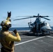 A flight deck crew member in yellow gear is signaling a military helicopter on the deck of a naval vessel at sea. The crew member is using hand signals to communicate with the helicopter crew, a standard procedure during flight deck operations. The helicopter, marked with the number '67', appears to be a U.S. Marine Corps UH-1Y Venom light utility helicopter.