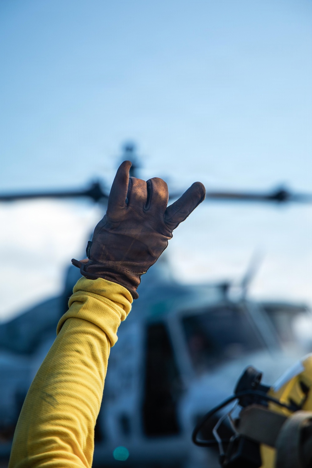 A person wearing a yellow long-sleeve shirt and brown leather work glove raises their hand in a signaling gesture, with index finger and pinky extended. In the blurred background, a military helicopter (likely a UH-1 Huey or similar rotary-wing aircraft) is visible on what appears to be a flight deck or landing pad. The image appears to depict a flight deck crew member directing or signaling aircraft operations.