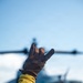 A person wearing a yellow long-sleeve shirt and brown leather work glove raises their hand in a signaling gesture, with index finger and pinky extended. In the blurred background, a military helicopter (likely a UH-1 Huey or similar rotary-wing aircraft) is visible on what appears to be a flight deck or landing pad. The image appears to depict a flight deck crew member directing or signaling aircraft operations.