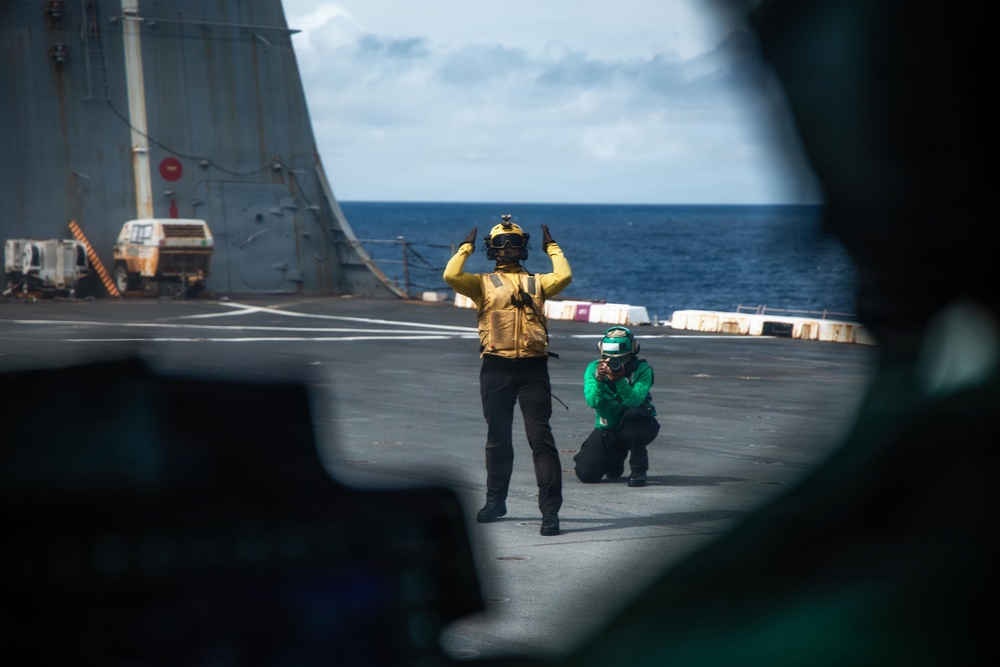 The image shows two naval personnel on the flight deck of a warship at sea. One person wearing a yellow vest and helmet is giving hand signals with both arms raised, while a second person in a green jersey and helmet crouches nearby, appearing to observe or photograph. The scene is viewed through what appears to be a cockpit window or aircraft frame in the foreground.